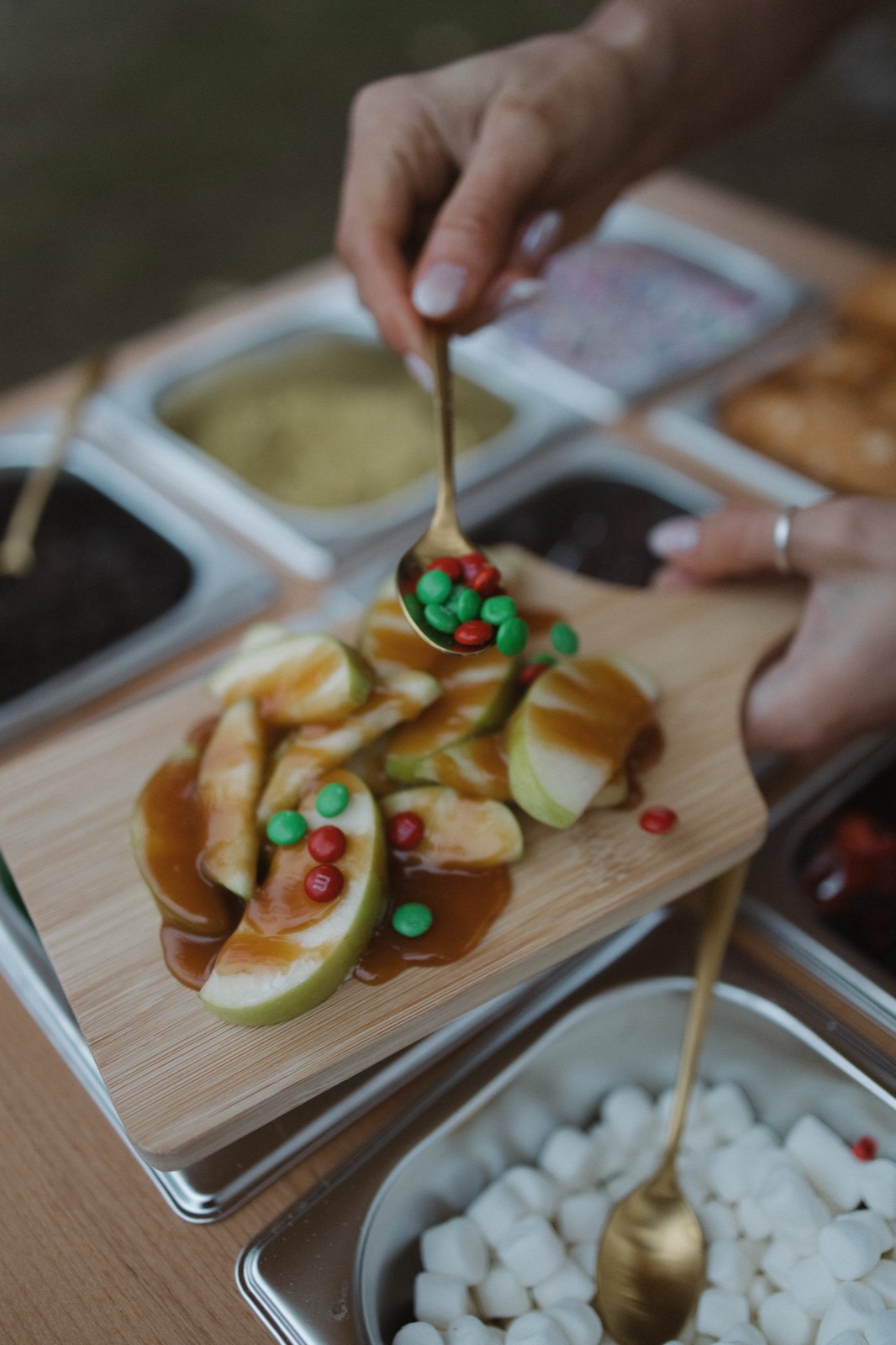 A spoon adding red and green candy pieces to caramel apple slices on a wooden board