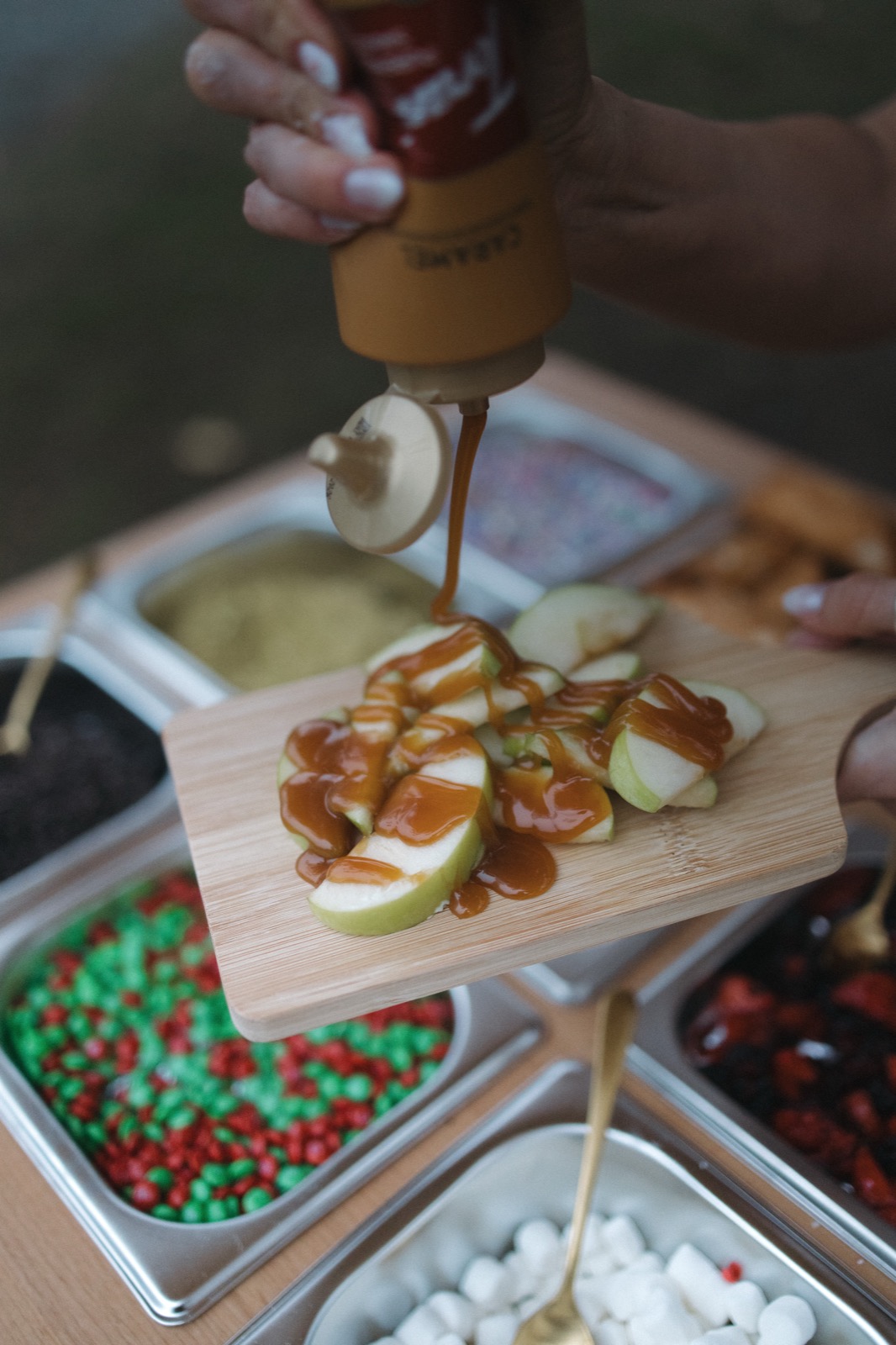 Caramel being drizzled over sliced green apples on a wooden serving board, with topping bar containers in the background