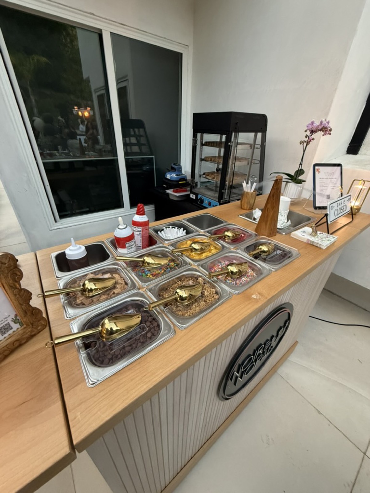 The full topping bar at a private event, with drizzles, dried fruit, nuts, and candy toppings in labeled tins