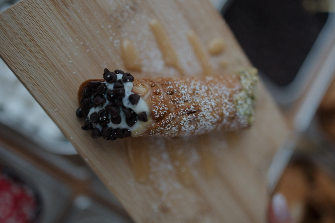 Close-up of a cannoli dipped in mini chocolate chips, with powdered sugar and caramel drizzle