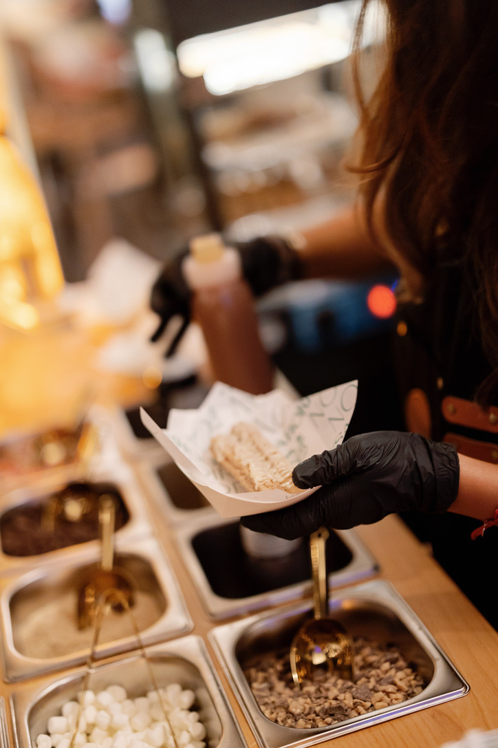 A staff member holding a warm churro over the topping bar, dipping sauce in hand