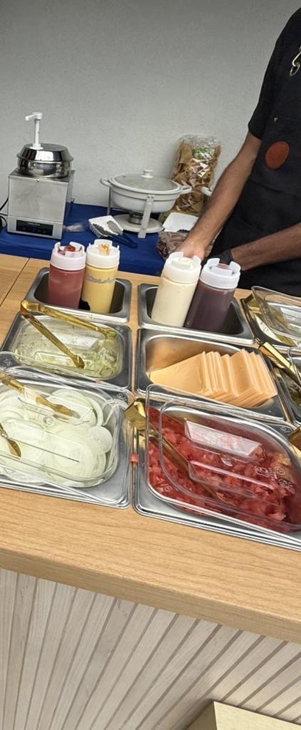 A savory topping bar with cheese, onions, diced tomatoes, and condiments, staff serving behind