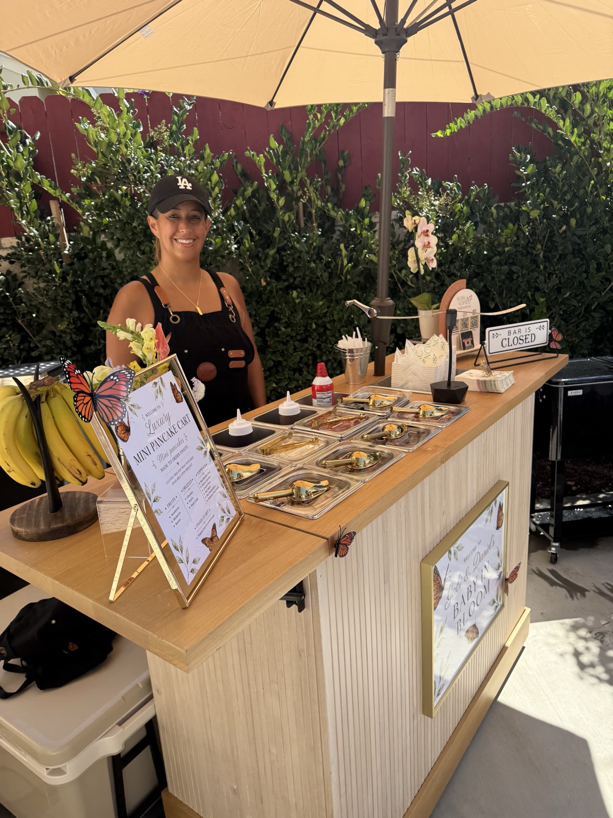 A staff member behind the styled Luxe Mini Pancake Stack cart, topping bar set up under an umbrella at a real event