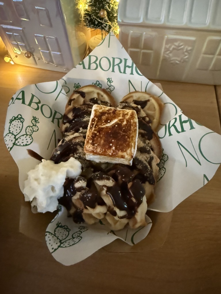 A finished topping presentation with chocolate drizzle, a toasted marshmallow, and whipped cream, served in Noir & Nopal branded paper