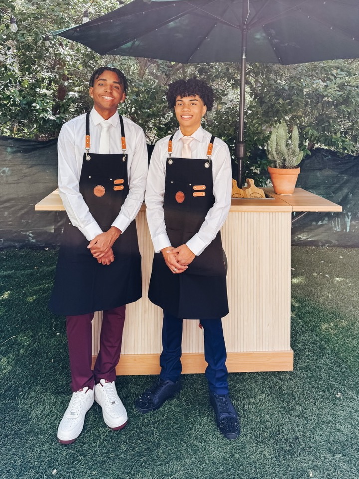 Two Noir & Nopal staff members in black aprons and white shirts standing at an event cart